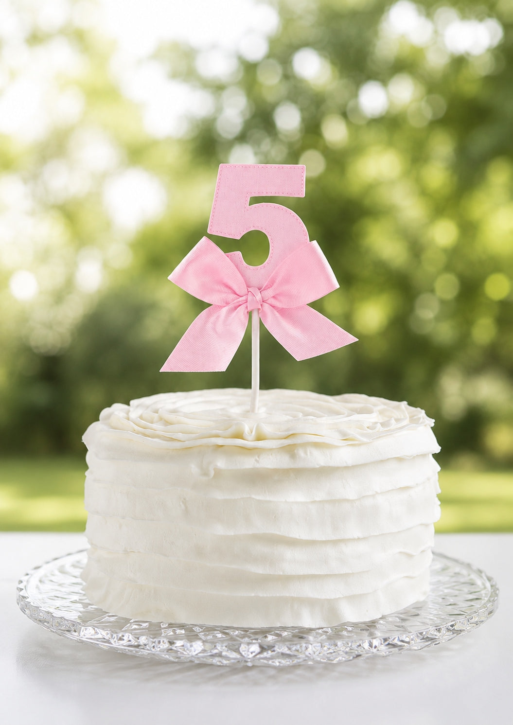 Handmade pink linen number five cake topper with a satin bow on a white buttercream cake on a glass stand