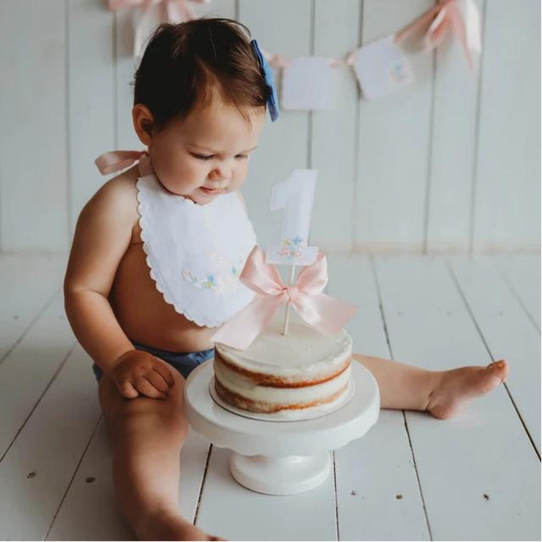 Baby in a handmade white linen first birthday bib reaching for a small smash cake on a white cake stand