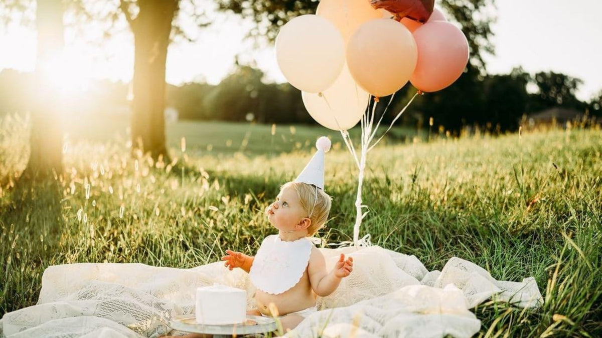 Baby in a handmade party hat and scalloped bib on a lace blanket at golden hour with a smash cake and peach balloons
