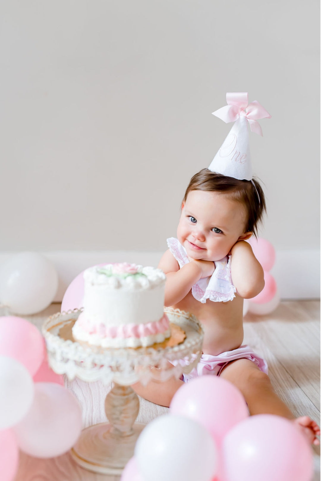 Baby in a handmade linen party hat beside a white smash cake surrounded by pink and white balloons
