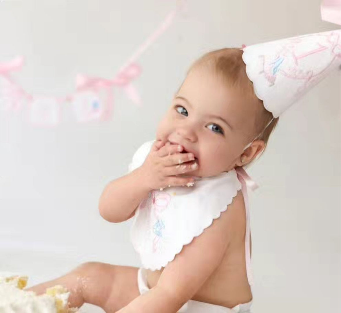 Close-up of a baby in a handmade scalloped bib and party hat tasting cake in front of a pink ONE banner